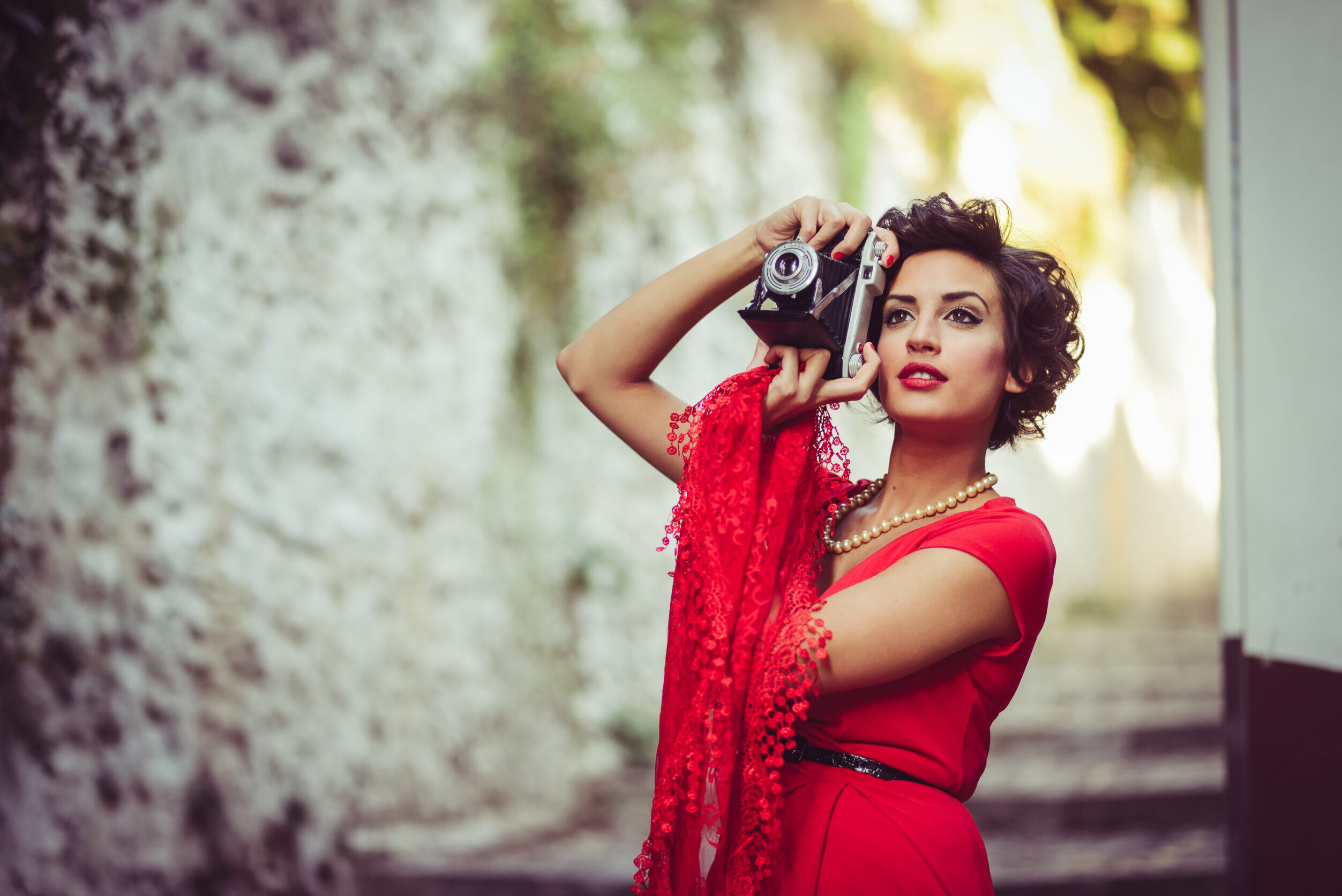 Portrait of a pretty woman, vintage style, in urban background, wearing a red dress taking photographs with a old camera
